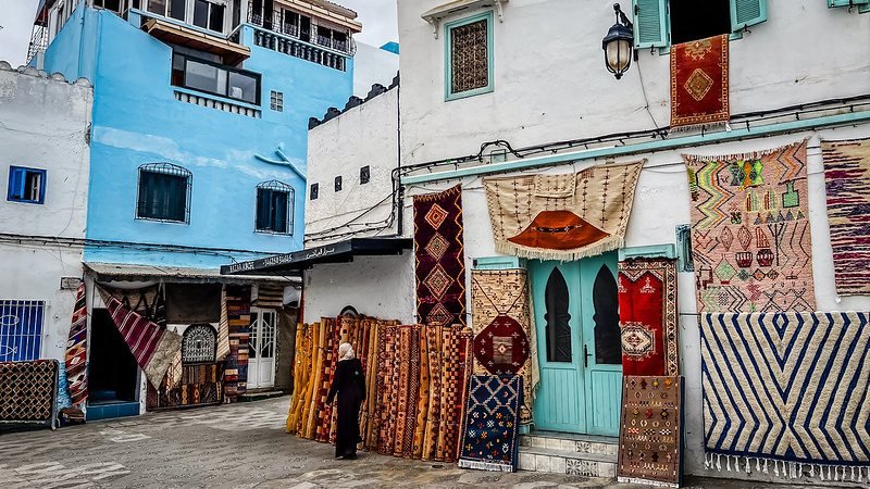 Carpets hung across a whitewashed lane in the Asilah medina, a woman walking past