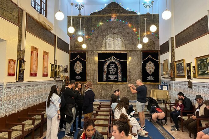 Interior of a synagogue in Tangier, its walls lined with Hebrew memorial plaques, visitors seated on the wooden benches